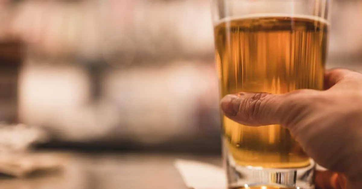 A close-up image of a hand holding a beer glass indoors on a table.